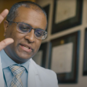 Indian man waves hand; he is wearing a blue shirt and a white physicians coat