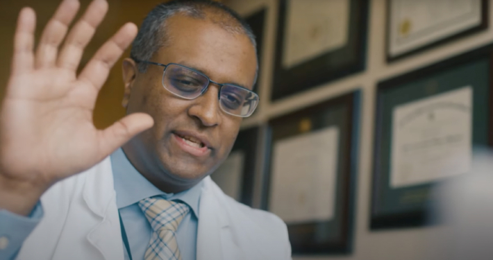 Indian man waves hand; he is wearing a blue shirt and a white physicians coat